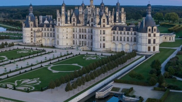 Ein prächtiges Schloss mit mehreren Türmen und einem symmetrischen Garten, darunter ein zentraler Brunnen und gepflegte Rasenflächen.
