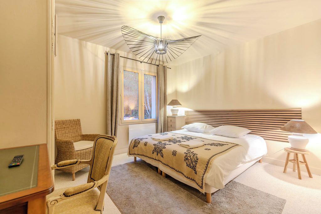Une chambre d’hôtel avec un lit, une chaise, un bureau avec une télécommande, une lampe et une fenêtre avec des rideaux.