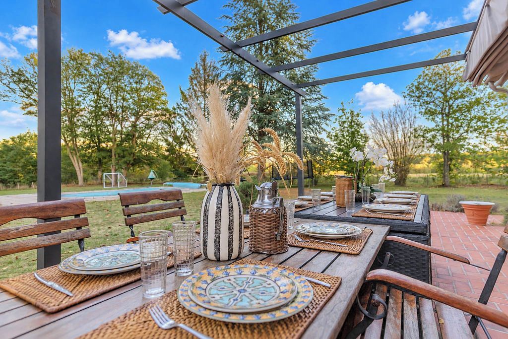 Un espace extérieur aménagé pour les repas, avec une table en bois dressée pour deux personnes, entourée de chaises, et offrant une vue sur une piscine et des arbres en arrière-plan.