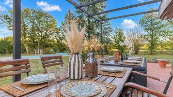 Un espace extérieur aménagé pour les repas, avec une table en bois dressée pour deux personnes, entourée de chaises, et offrant une vue sur une piscine et des arbres en arrière-plan.