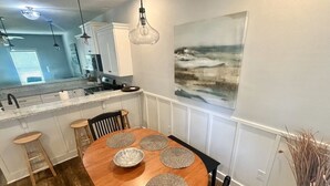 Een eetgedeelte met een houten tafel, vier placemats en een kom. Twee barkrukken en een stoel staan er. Achtergrond is een keuken met witte kasten en een hangende lamp.