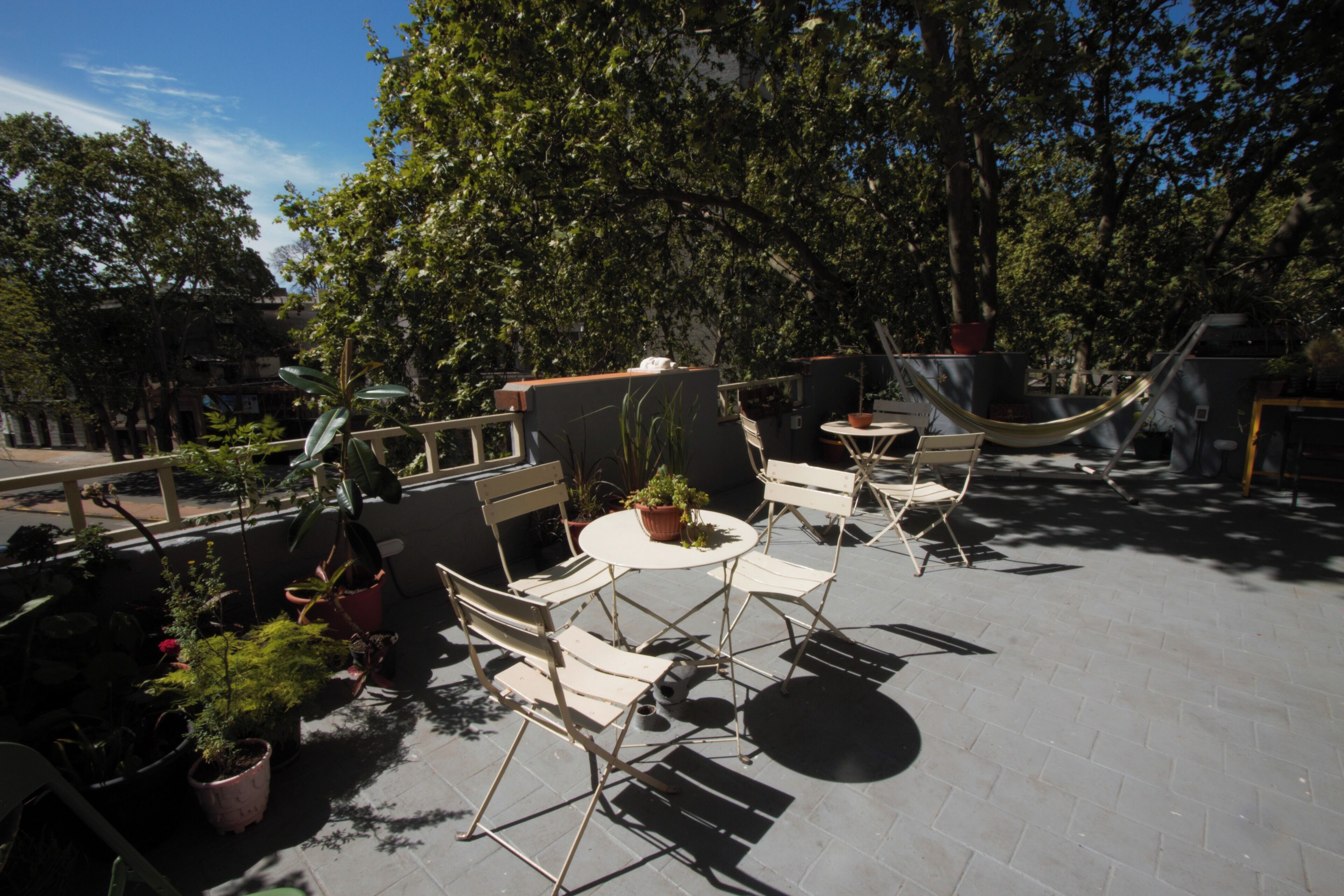 An outdoor patio with a round table and four chairs, surrounded by potted plants and a hammock.