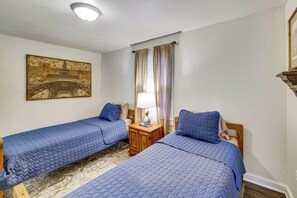 A room with two single beds, a nightstand with a lamp, a painting on the wall, and a window with curtains.