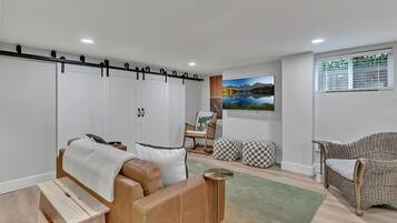 A modern living room with a brown leather sofa, a wooden bench, a wicker chair, and a checkered ottoman. There is a painting on the wall and a window with blinds.