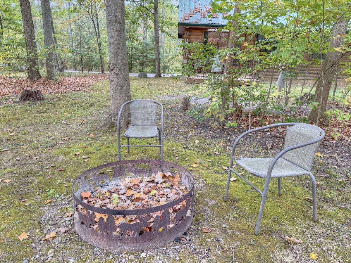 Un espace de détente extérieur de style rustique, avec un foyer en métal et deux chaises en métal, entouré d’arbres et agrémenté d’un chalet en bois en arrière-plan.