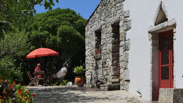 Un edificio de piedra con una puerta roja, un patio con una mesa y sillas, y un paraguas rojo.