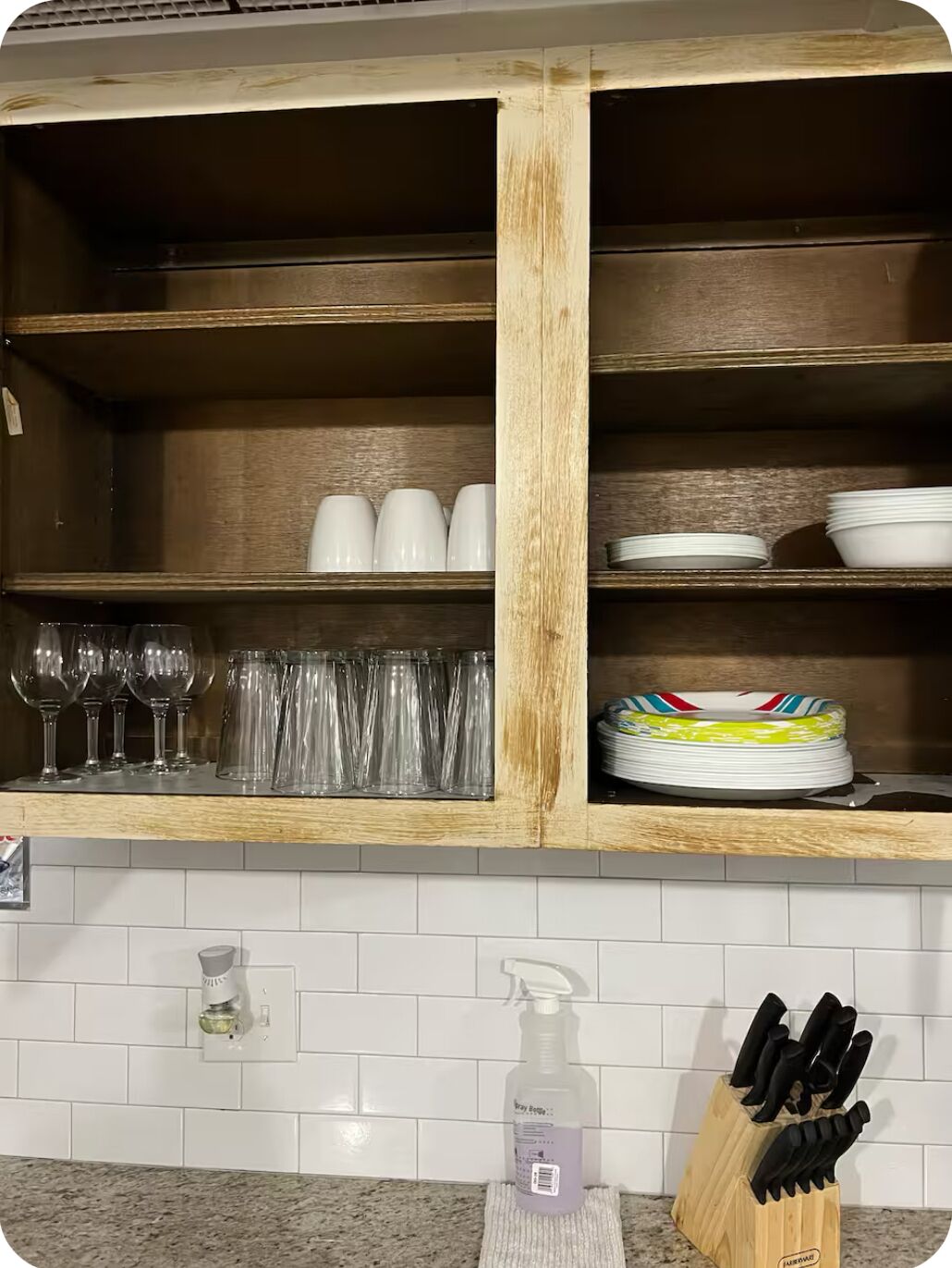 Cocina con armarios de madera, un lavamanos y una encimera. Hay copas de vino, tazas y platos guardados en los armarios. En la encimera se encuentran un pulverizador y un bloque de cuchillos.
