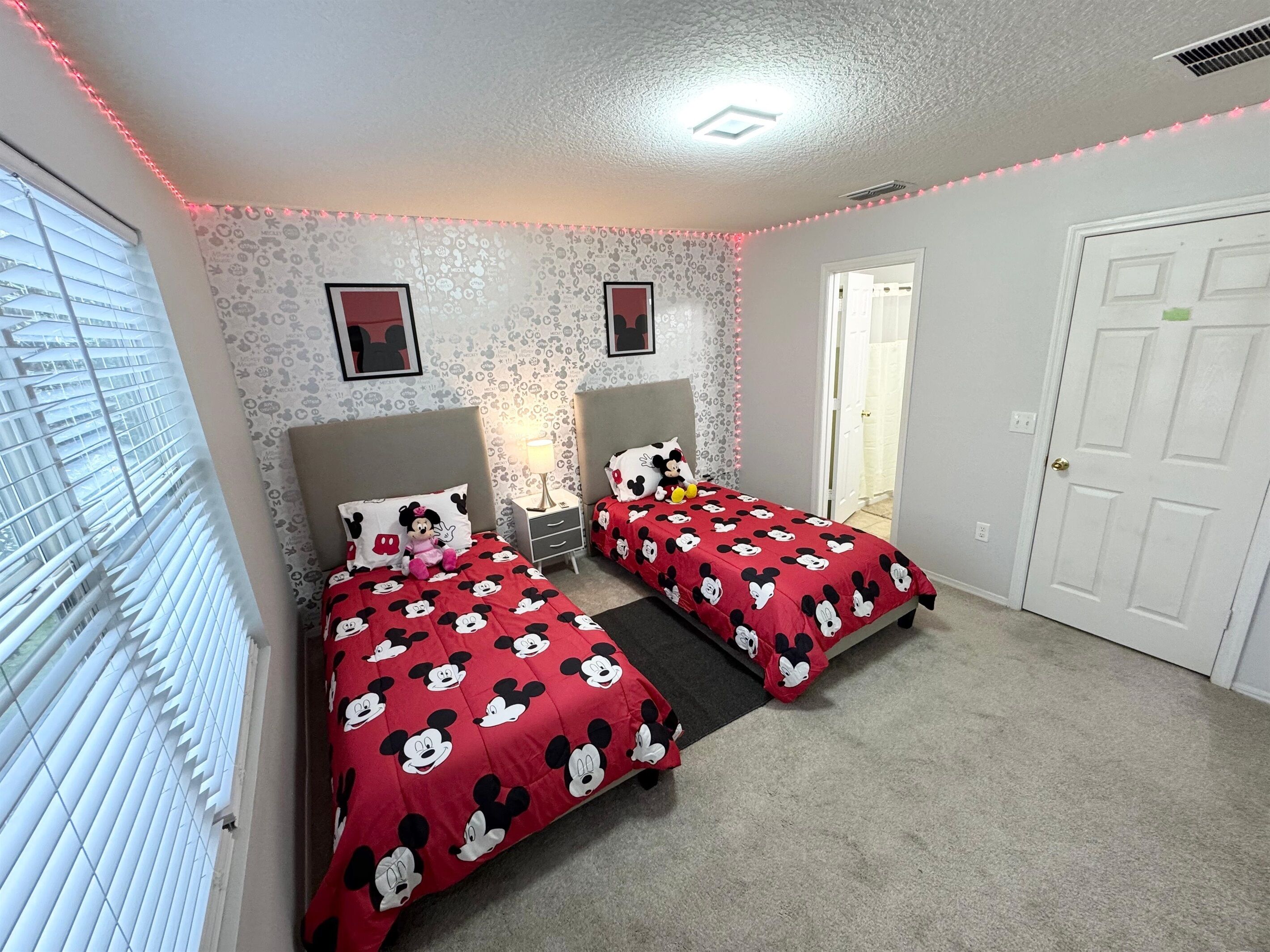 Een slaapkamer met twee bedden, elk met een Mickey Mouse sprei, een nachtkastje met een lamp en een raam met jaloezieën.