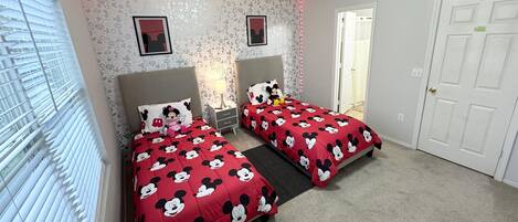 Een slaapkamer met twee bedden, elk met een Mickey Mouse sprei, een nachtkastje met een lamp en een raam met jaloezieën.