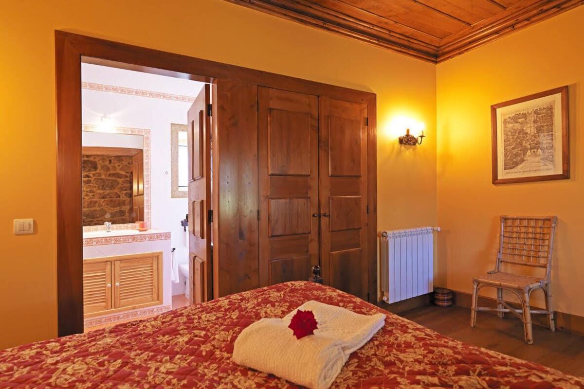 A bedroom with a wooden door, a bed with a red and white patterned bedspread, a chair, and a framed picture on the wall.