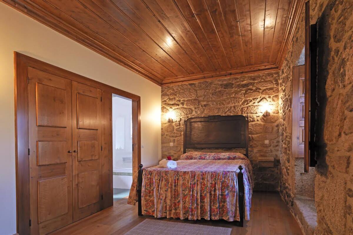 A bedroom with a wooden ceiling, stone walls, a bed with floral bedding, and a nightstand.