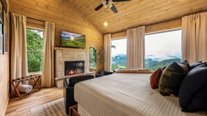 Une chambre avec une cheminée, un lit avec des oreillers, un ventilateur de plafond et de grandes fenêtres avec des rideaux.