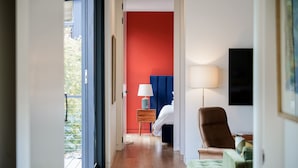 Quarto de hotel com parede vermelha de destaque, cama com roupa de cama branca, cabeceira azul, mesa de cabeceira com abajur e uma cadeira.