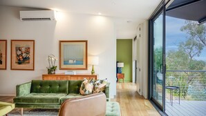Uma sala de estar moderna com um sofá verde, uma mesa de centro e uma pintura na parede. Há uma janela com vista para árvores e uma varanda.