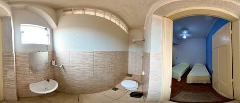 A bathroom with a sink, toilet, and shower area, featuring a curved ceiling and a view into another room with beds.
