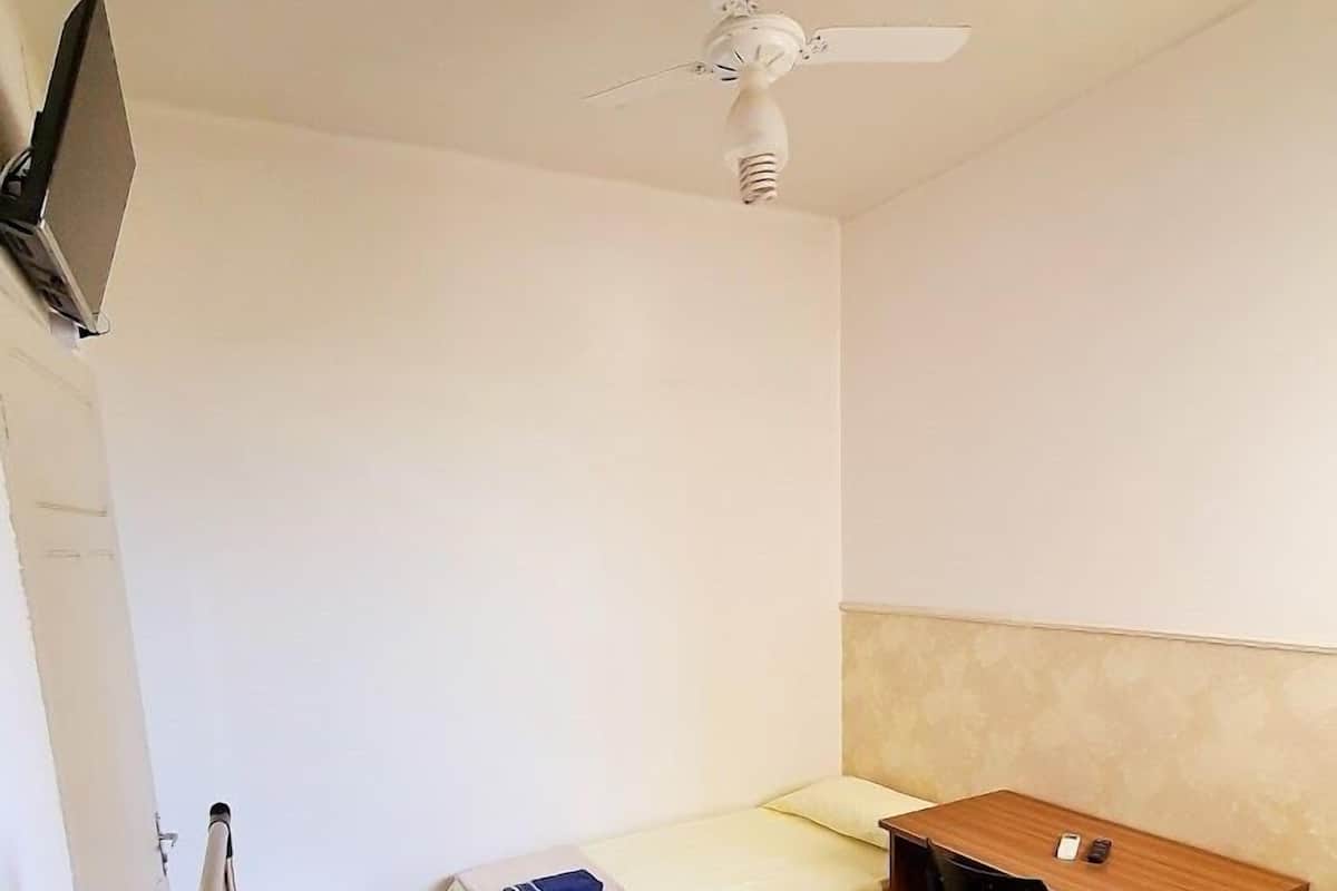 A small, clean room with two single beds, a wooden desk, a ceiling fan, and a television mounted on the wall.