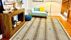A living room with a blue sofa, a patterned rug, a wooden console table with books and a plant, and a cityscape painting. - Family Retreat - Stunning 5BR-2BA with Parking/ Near Savin Hill Station (Boston)