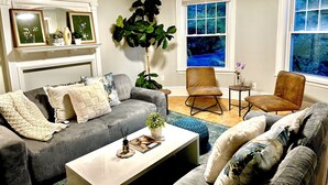 A living room with a grey sofa, a brown armchair, and a white coffee table. There is a large plant in the corner and a window with a view of trees. - Family Retreat - Stunning 5BR-2BA with Parking/ Near Savin Hill Station (Boston)