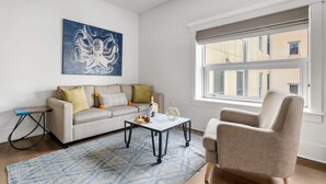 A living room with a sofa, a coffee table, a chair, a painting, and a window with a view of buildings. - The Quad at 493 - 4 Suites - Right on King! (Charleston)