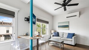 A modern living room with a white sofa, a glass dining table, and a clear chair. There is a ceiling fan, a window with a view of buildings, and an air conditioning unit on the wall. - The Quad at 493 - 4 Suites - Right on King! (Charleston)