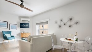 A modern living room with a sofa, a blue armchair, and a dining area with clear chairs and a table set with a bottle, glasses, and a fruit platter.