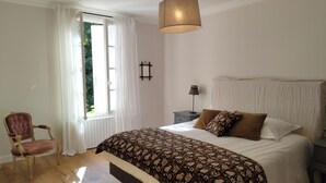 A bedroom with a large bed, a chair, a nightstand, a lamp, and a window with curtains.