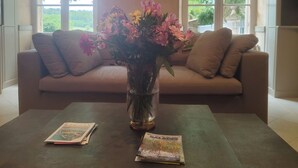 A living room with a beige sofa, a coffee table, and a vase of flowers. There are two windows with a view of greenery outside.