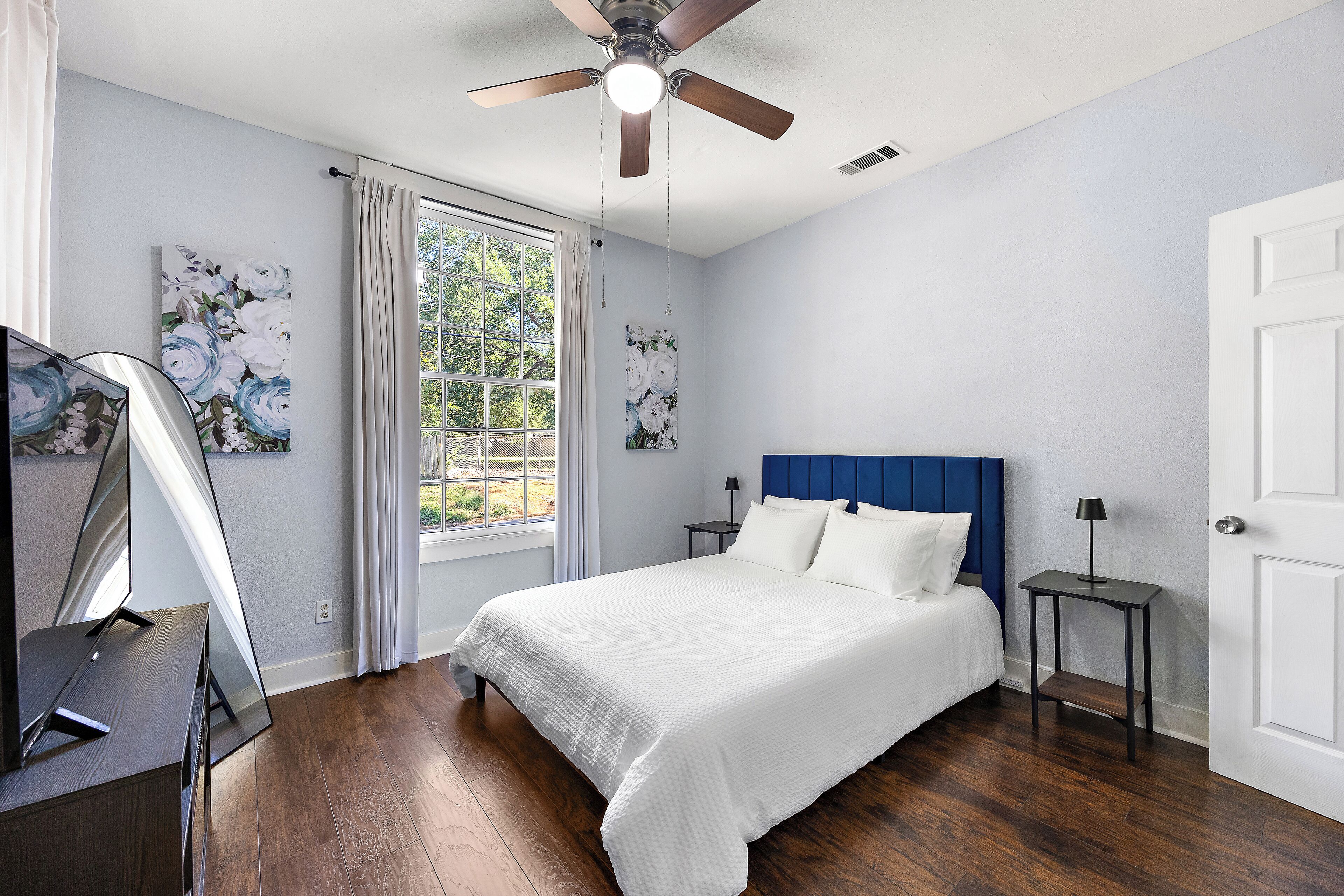 A bedroom with a bed, a ceiling fan, a window with curtains, and artwork on the walls.