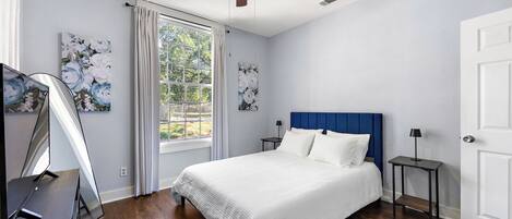 A bedroom with a bed, a ceiling fan, a window with curtains, and artwork on the walls.