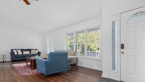 A spacious living room with a blue sofa, a blue armchair, and a wooden coffee table. There are large windows with blinds, a white door, and a ceiling fan. - Near Newport, Beaches, Golf & Navy Base (Middletown)