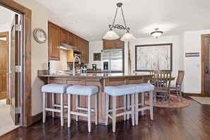 A kitchen with wooden cabinets, a stainless steel refrigerator, a bar with stools, and a hanging light fixture.