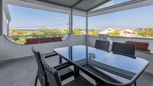 En balkon med et glasbord og stole, plantekasser og udsigt til huse og grøn bevoksning.
