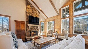 A cozy log cabin interior with a stone fireplace, wooden walls, and large windows offering a view of the outdoors.