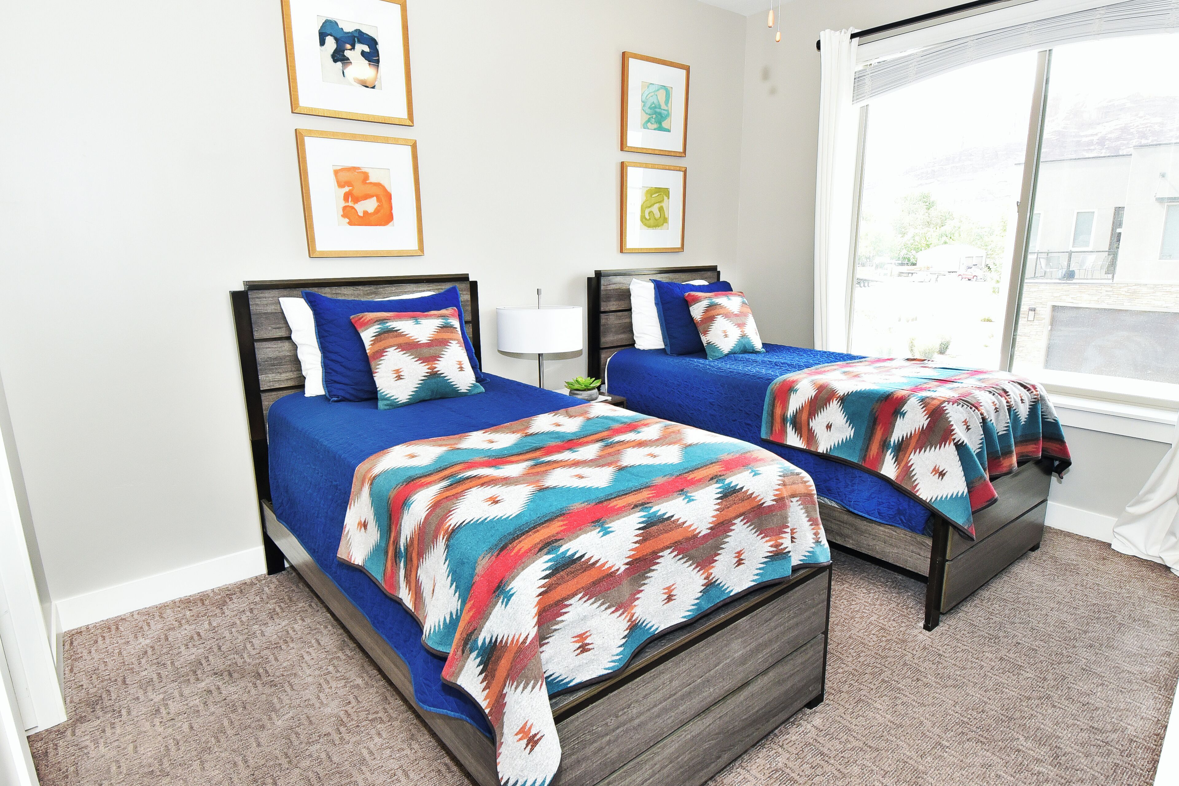 Una habitación con dos camas, cada una con cubrecama azul y manta colorida. Hay cuadros en la pared y una ventana con vistas al exterior.