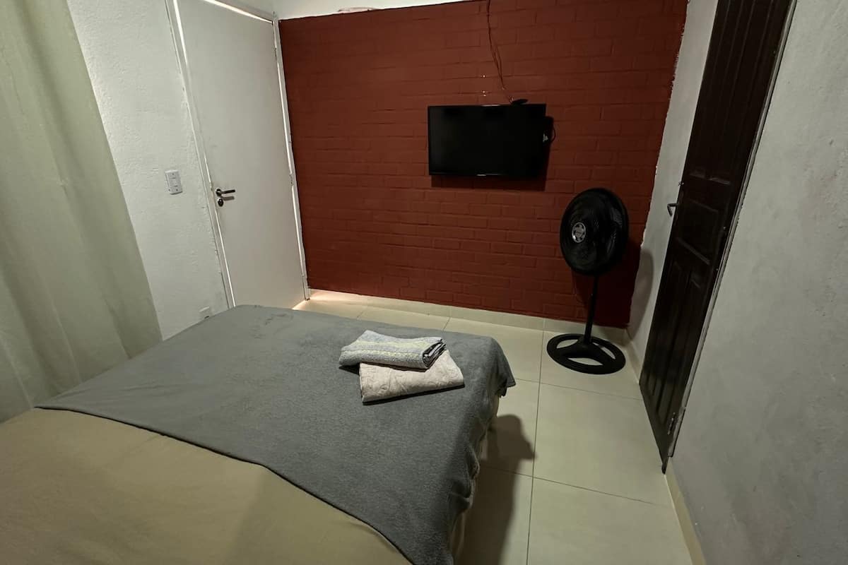 A small, neatly arranged bedroom with a bed, a flat-screen TV mounted on a brick wall, and a standing fan.