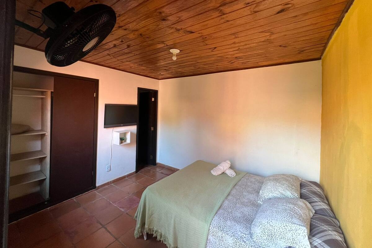 A bedroom with a wooden ceiling, a bed with patterned bedding, a TV mounted on the wall, and a small shelf with a plant.