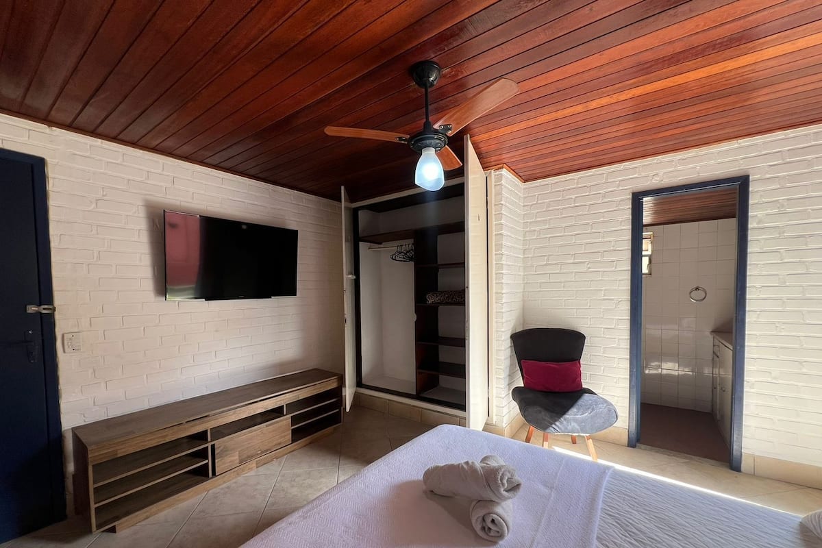 A bedroom with a wooden ceiling, a TV mounted on the wall, a bench, a bed with a rolled towel, a small table with a chair, and a shower area.