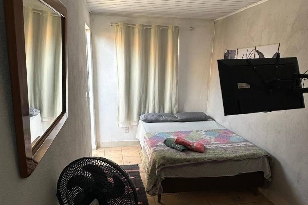 A small, well-lit room with a bed, a standing fan, a TV mounted on the wall, and a window with curtains.
