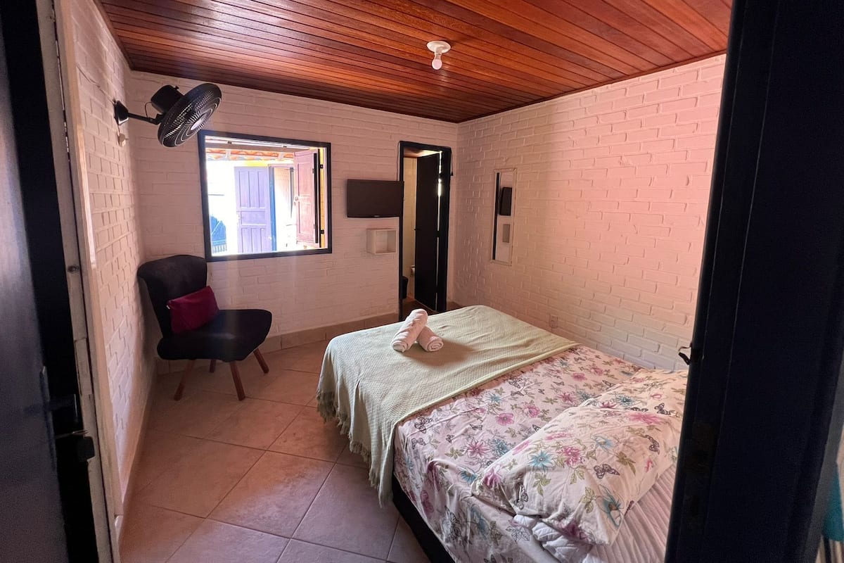 A small, cozy room with a bed, a chair, a TV, and a window.