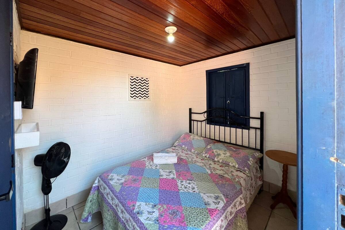 A small bedroom with a wooden ceiling, a bed with a colorful quilt, a small table, and a window with a dark blue shutter.