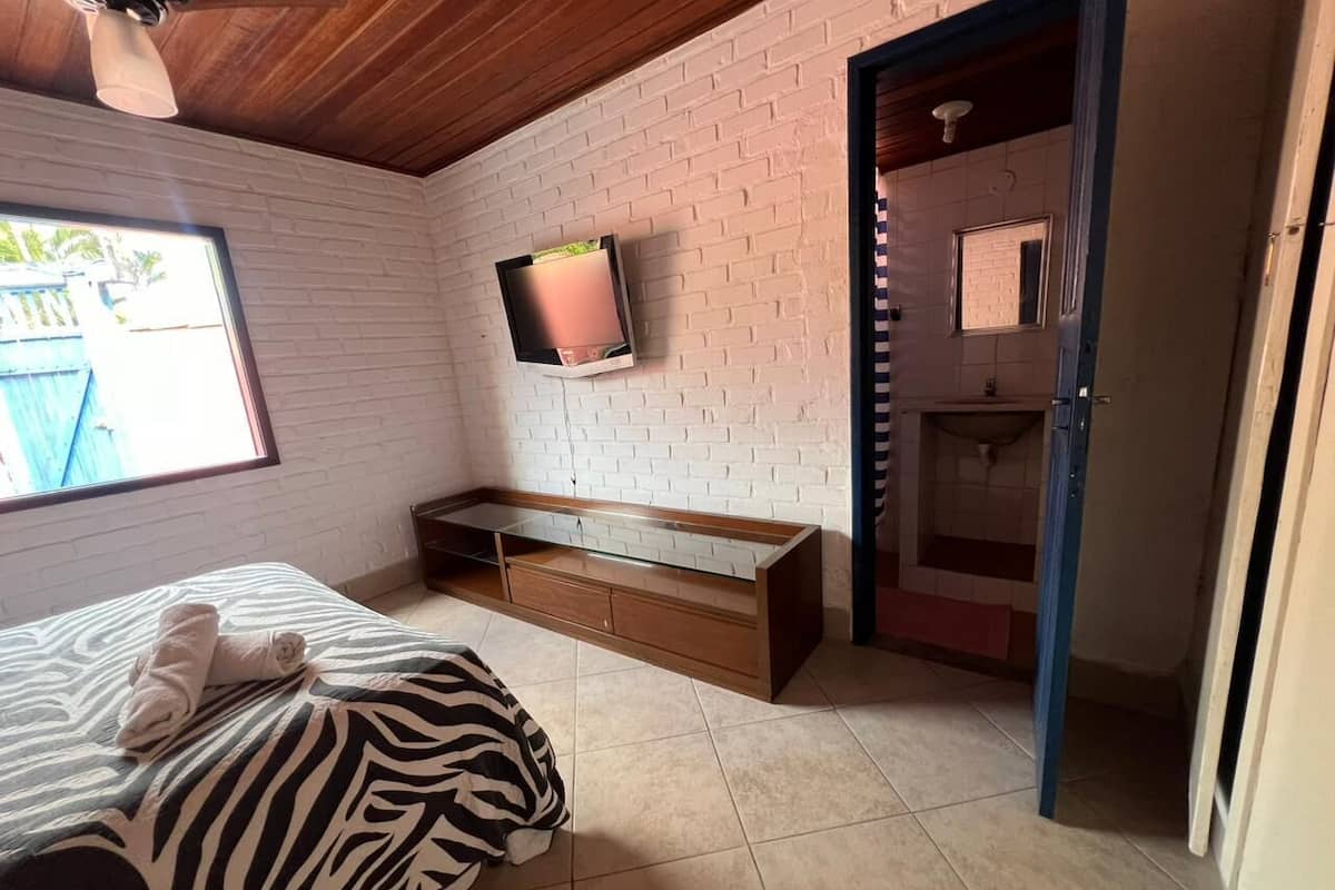 A bedroom with a bed, a zebra-patterned blanket, a wooden bench, a TV mounted on a brick wall, and a bathroom visible through an open door.