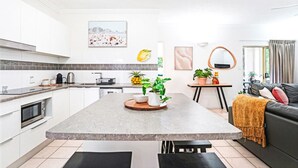 Una cocina moderna con armarios blancos, encimera de granito y una isla central con plantas. Hay una zona de comedor con un sofá negro y una mesa de centro.
