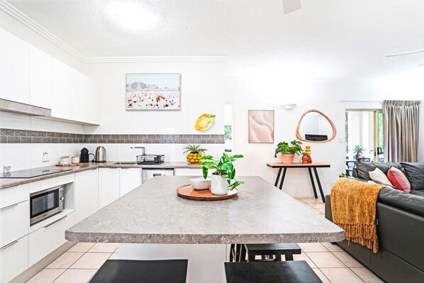 A modern kitchen with white cabinets, a granite countertop, and a central island with plants. There is a dining area with a black sofa and a coffee table.