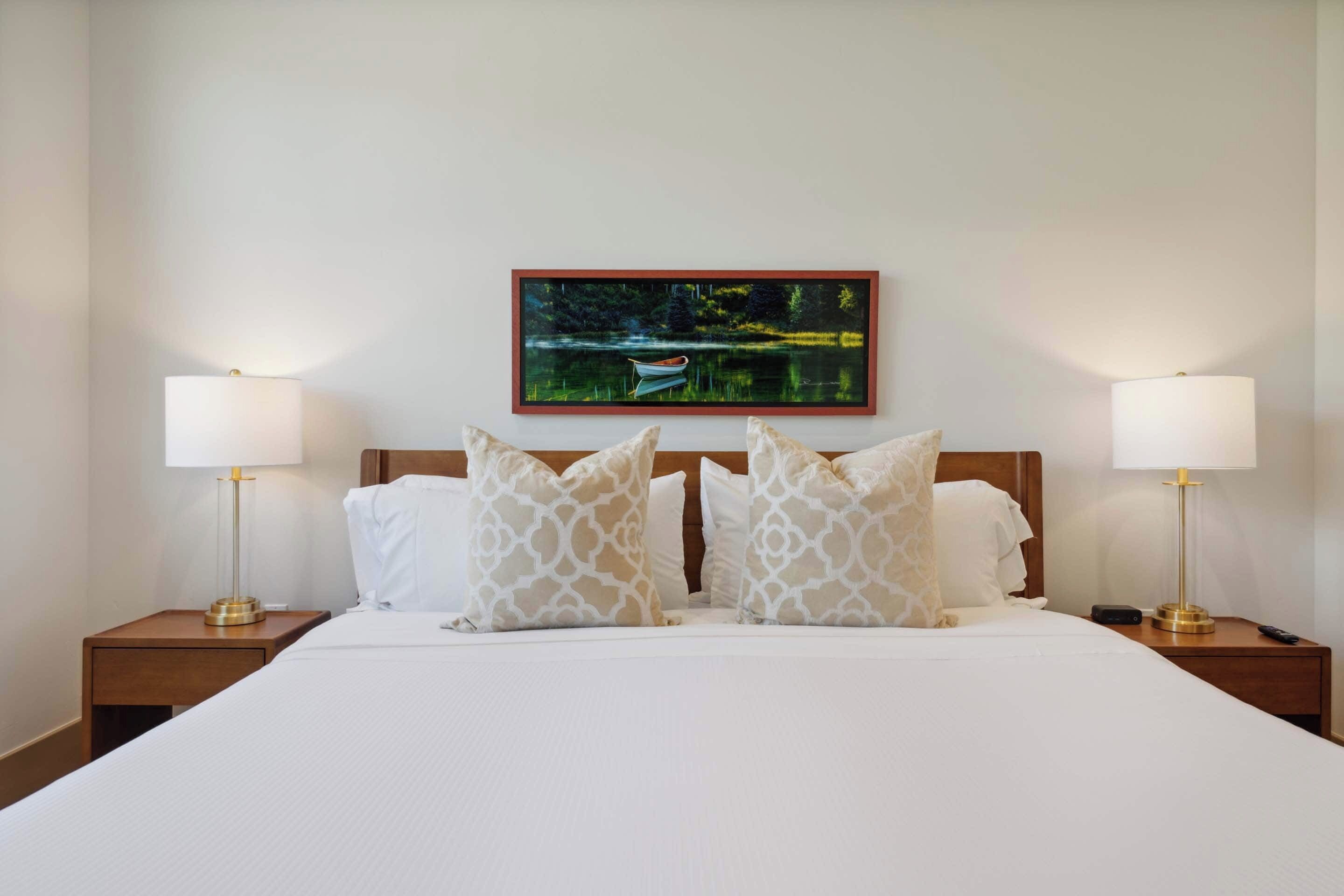 A hotel room with a large bed, two bedside lamps, a headboard, and a framed picture of a lake on the wall.