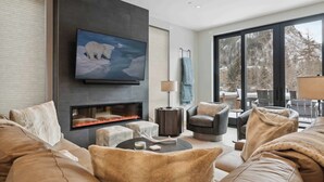 A modern living room with a fireplace, comfortable seating, a large TV, and a view of snowy mountains. - One Riverfront Townhome 05 (Avon)