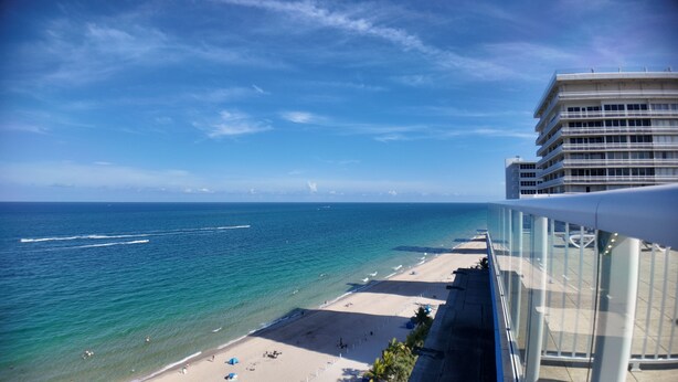 A beach view from a balcony with a clear sky, ocean, and a hotel building in the background. - Two Bedroom Penthouse with Large Balcony overlooking the Ocean spectacularview (Fort Lauderdale)