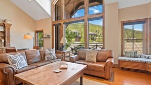 A spacious living room with a high ceiling, large windows offering a view of a mountainous landscape, a wooden coffee table, and leather sofas.