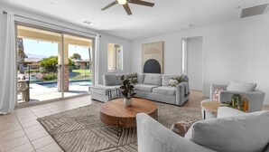 A spacious living room with a grey sofa, a coffee table, and a view of a swimming pool through large glass doors. - Agave House ~ Free Pool and Spa 🏊‍♀️ Heat  🐕 Pets Welcome !  Onsite ⛳️🎾 (Indio)