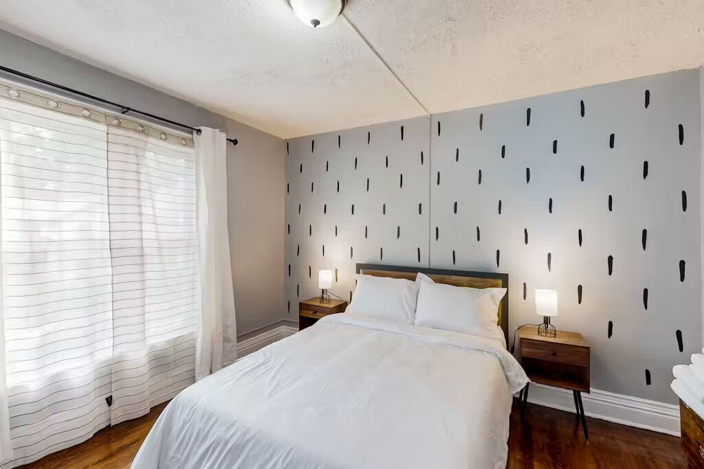 A bedroom with a bed, two bedside tables, a nightstand, a window with blinds, and a wall with a geometric pattern.