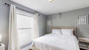 Un dormitorio con cama, mesita de noche, ventana con cortinas y cuadros en la pared.